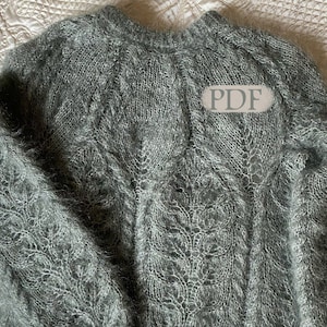 Puede incluir: Un suéter gris tejido a mano con un delicado patrón calado. El suéter tiene mangas largas y cuello redondo. La palabra "PDF" es visible en el suéter. El hilo parece suave y ligeramente difuso.
