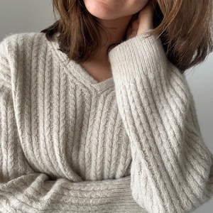 May include: A beige cable-knit sweater with a V-neck. The sweater has a textured pattern and long sleeves. The sleeves are cuffed at the wrists. The sweater appears to be made of a soft, warm material.