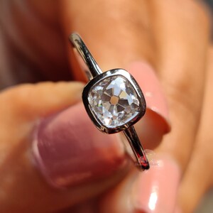 May include: A silver ring with a square, clear gemstone. The gemstone is set in a bezel setting and has multiple facets that reflect light. The ring is held up by a person with pink nail polish.