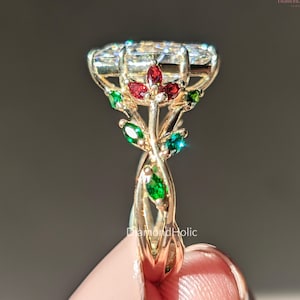 Op de afbeelding: Een gouden ring met een grote, heldere edelsteen en een bloemmotief. De ring heeft rode en groene accentstenen, met een gedraaide band. De ring is geplaatst tegen een neutrale achtergrond.