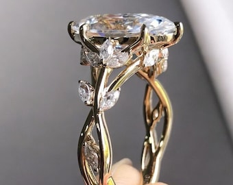 May include: A gold ring with a large, oval-cut diamond as the centerpiece. The band features a delicate, intertwined vine design with small, leaf-shaped diamonds. The ring is held up by a hand, showcasing the intricate details of the jewelry.