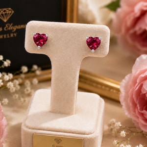May include: A pair of heart-shaped stud earrings featuring vibrant red gemstones set in silver-toned metal. The earrings are displayed on a beige velvet T-bar stand with a gold-toned plaque that reads 6 mm Heart Studs.