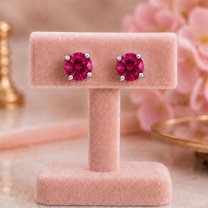 May include: A pair of round, brilliant-cut fuchsia pink stud earrings set in silver-toned metal. The earrings are displayed on a rectangular, light pink velvet T-bar stand, showcasing the vibrant color and faceted texture of the stones.