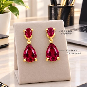May include: A pair of gold-toned drop earrings featuring two faceted, pear-shaped red stones each. The top stone measures 8 x 6 mm, and the bottom stone measures 10 x 14 mm. The earrings are displayed on a light beige, triangular-shaped jewelry stand.