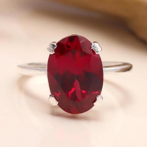 May include: A silver ring featuring a large, oval-cut, deep red gemstone. The gemstone is the focal point, set with four prongs. The band is a simple, polished silver. The ring is displayed on a light-colored surface.