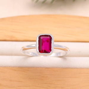 Peut inclure: Bague en argent avec une pierre précieuse rectangulaire rouge foncé. La pierre est sertie dans un sertissage à lunette, et le bracelet est fin et poli. La bague est présentée sur une surface claire, avec un fond en bois.