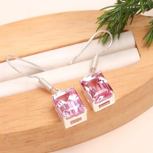 May include: A pair of silver drop earrings with rectangular, light pink gemstones. The earrings are displayed on a wooden surface with a white insert, and a sprig of greenery is visible in the background.