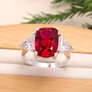 Può includere: Un anello d'argento con una grande gemma rossa rettangolare vibrante come elemento centrale, affiancata da due gemme chiare triangolari più piccole. L'anello è posto su uno sfondo chiaro.