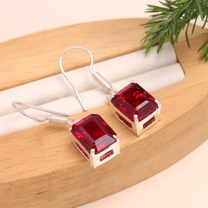 May include: Silver earrings featuring large, rectangular, deep red gemstones. The gemstones are set in a silver frame and have a faceted appearance. The earrings are displayed on a wooden surface with a white interior.