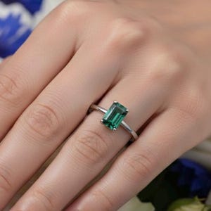 Peut inclure: Une bague argentée ornée d'une pierre précieuse verte de taille émeraude rectangulaire, fixée par quatre griffes. L'anneau est fin et poli, mettant en valeur une pierre verte claire et éclatante posée sur le doigt devant un fond floral.