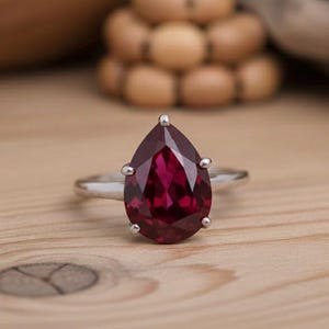Peut inclure: Une bague en argent ornée d'une grosse pierre précieuse rouge foncé en forme de poire. La pierre est facettée et sertie de quatre griffes. La bague est posée sur une surface en bois brun clair.