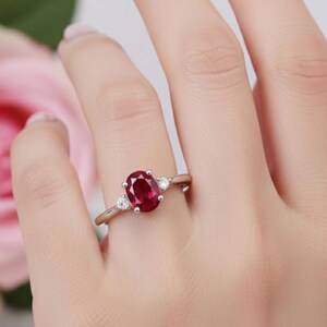 May include: A silver ring featuring a large oval-cut ruby gemstone, flanked by two smaller, round, clear gemstones. The ring is set against a soft background with a pink rose.