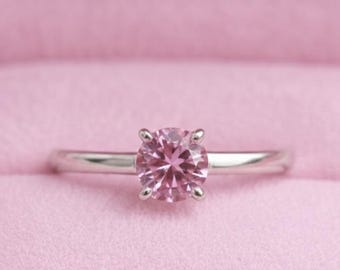 May include: A silver ring featuring a round, pale pink gemstone. The ring is set in a four-prong setting and is presented in a pink ring box. The ring's band is slender and polished.