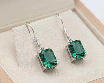 May include: A pair of emerald-cut green gemstone earrings with silver-colored metal settings and hooks. The earrings are displayed inside a light-colored, wooden jewelry box with a cream-colored interior.