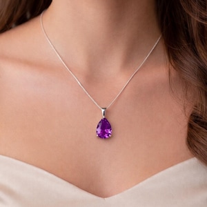May include: A silver necklace featuring a pear-shaped, faceted purple gemstone pendant. The pendant is suspended from a delicate silver chain that rests against the collarbone, showcasing a clear, vibrant violet hue in the stone.
