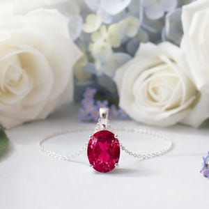 May include: A silver necklace featuring a large, oval ruby pendant. The vibrant red gemstone is set in a silver mount and hangs from a fine chain. The background includes white roses and other flowers, creating a soft, romantic scene.