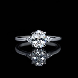 Könnte beinhalten: Ein silberner Verlobungsring mit einem oval geschliffenen Diamanten als Mittelstein und zwei kleineren Seitensteinen. Der Ring ist vor einem schwarzen Hintergrund abgebildet, wobei sich der Ring spiegelt.