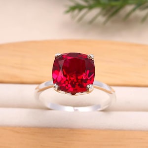 Può includere: Un anello d'argento con una grande gemma rossa brillante a taglio quadrato. La gemma è il punto focale, incastonata in una classica montatura a quattro griffe. La fascia dell'anello è in argento lucido semplice, creando un contrasto sorprendente con la pietra rossa.