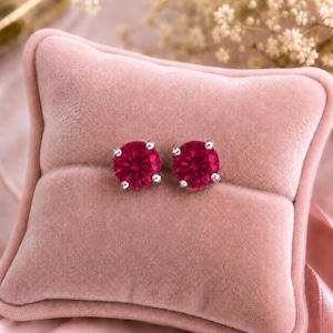 Puede incluir: Un par de pendientes de botón con piedras preciosas rojas de corte redondo engastadas en garras de metal plateado. Los pendientes se muestran uno al lado del otro, destacando la superficie facetada de las piedras y el diseño de engaste.