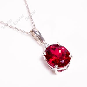 May include: A silver pendant necklace featuring a large, oval-cut, deep red gemstone. The gemstone is set in a silver bezel with four prongs and a small accent stone above. The chain is delicate.