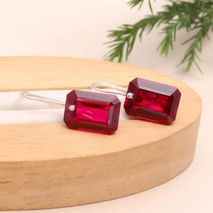 May include: A pair of ruby-red rectangular earrings with silver hooks. The earrings have a faceted design, catching the light. The earrings are displayed on a wooden surface, with green foliage in the background. The earrings are a jewelry item.