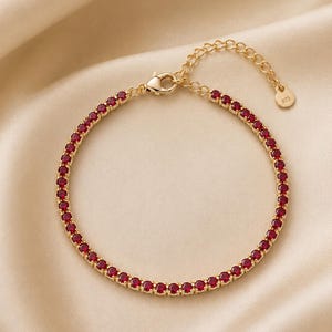 Op de afbeelding: Een goudkleurige tennisarmband met een doorlopende rij ronde, dieprode stenen. De armband is voorzien van een karabijnsluiting, een verstelbare verlengketting en een kleine ronde bedel met de inscriptie 925 voor metaalzuiverheid.