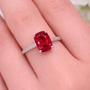 Peut inclure: Une bague en argent ornée d'une grosse pierre rouge vif de forme rectangulaire. La monture est ornée de petites pierres scintillantes. La bague est présentée sur une main, mettant en valeur le design et la couleur du bijou.