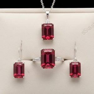 May include: A jewelry set featuring a pendant necklace, ring, and earrings. Each piece has a rectangular, deep red gemstone set in a silver-colored metal. The ring has two small, clear stones on either side of the main stone.