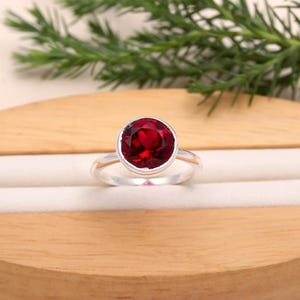 Könnte beinhalten: Ein silberner Ring mit einem großen, runden, tiefroten Edelstein. Der Edelstein ist facettiert und in einer Lünettenfassung gefasst. Das Band ist glatt und poliert. Der Ring wird auf einer hellen Oberfläche mit einem Holz- und Grünpflanzenhintergrund präsentiert.