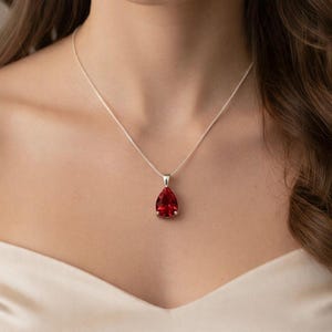 Op de afbeelding: Een zilveren ketting met een druppelvormige, helderrode edelsteen hanger. De hanger is gezet in een zilveren zetting en hangt aan een fijne zilveren ketting. De ketting wordt getoond tegen een neutrale achtergrond.