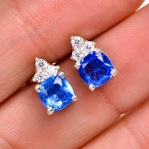 Könnte beinhalten: Ein Paar silberne Ohrstecker mit einem blauen Edelstein und kleineren weißen Edelsteinen an den Rändern.