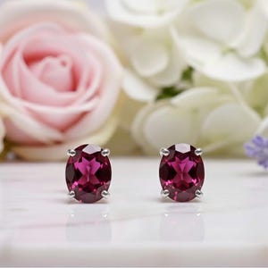 May include: A pair of oval-shaped, deep magenta gemstone stud earrings. The earrings are set in silver and are displayed on a white surface, with a blurred background of pink roses and white flowers.