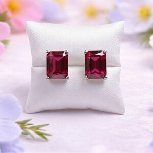 May include: Silver stud earrings, each featuring a rectangular, deep red gemstone. The earrings are presented on a white cushion, with a backdrop of soft-focus pink, purple, and white flowers.