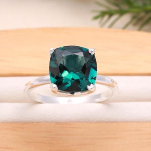 Op de afbeelding: Een zilveren ring met een grote, vierkant geslepen, smaragdgroene edelsteen. De gefacetteerde edelsteen reflecteert licht. De ringband is van gepolijst zilver. De zetting heeft vier klauwen.