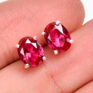 Peut inclure: Une paire de boucles d'oreilles à clous en forme d'ovale, avec des pierres précieuses rouge rubis. Chaque boucle d'oreille présente une pierre précieuse vibrante et facettée, sertie dans un sertissage à griffes en métal argenté.
