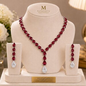 May include: A jewelry set featuring a necklace and two drop earrings displayed on a beige stand. The necklace has a Y-shaped design with pear-cut deep red stones and a single clear pear-cut stone at the bottom. Each earring has three red stones.