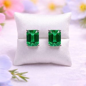 Puede incluir: Un par de pendientes de botón con piedras preciosas verdes esmeralda de corte rectangular. Los pendientes están engarzados en un metal plateado y se muestran sobre un cojín blanco. Flores rosas, blancas y azules en el fondo.