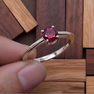 Könnte beinhalten: Ein silberfarbener Ring mit einem einzelnen, rund geschliffenen, tiefroten Edelstein in einer Vier-Krappen-Fassung. Die Ringschiene ist glatt und poliert und zeigt ein schlichtes Design, das die leuchtende Farbe und facettierte Oberfläche betont.