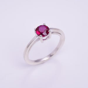 May include: A silver ring with a round, deep red gemstone. The gemstone is held in place by four prongs, and the band is plain, polished silver. The ring's design is classic, suitable for various occasions.