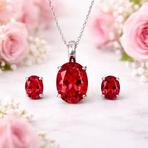 Puede incluir: Un conjunto de joyas con un collar con colgante y pendientes a juego. El colgante y los pendientes muestran grandes piedras preciosas rojas de corte ovalado engastadas en metal plateado. El collar tiene una cadena delicada.