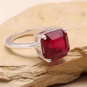 May include: A silver ring featuring a large, square, deep red gemstone. The gemstone is the focal point, set in a simple silver band. The ring is displayed on a light brown, textured surface.