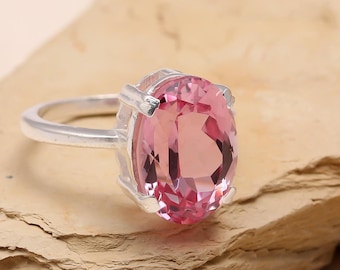 May include: A silver ring featuring a large, oval-cut pink gemstone. The gemstone is the focal point, set in a simple, elegant silver band. The ring is displayed on a textured, light brown surface, highlighting its color and design.