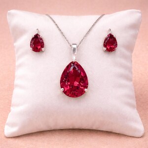 Può includere: Un set di gioielli con una collana d'argento con un grande pendente a forma di goccia di pietra rossa e orecchini abbinati. Il set è esposto su un cuscino di raso bianco su uno sfondo rosa tenue.