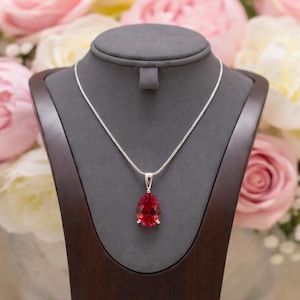 May include: A silver necklace with a pear-shaped, red gemstone pendant. The pendant is displayed on a dark wooden jewelry stand against a backdrop of pink and white flowers. The chain is delicate and the gemstone is faceted.