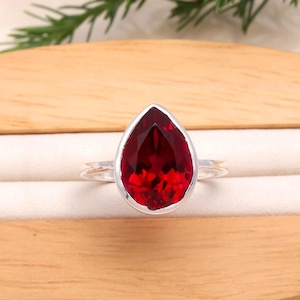 Peut inclure: Bague en argent ornée d'une grosse pierre rouge en forme de goutte d'eau. La pierre précieuse facettée est sertie dans un anneau en argent poli. La bague est présentée sur une surface claire, mettant en valeur sa couleur et son design.