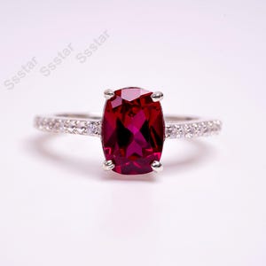 Peut inclure: Une bague en argent avec une grande pierre précieuse rouge de forme ovale entourée de petites pierres précieuses blanches.