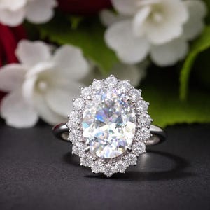 Könnte beinhalten: Ein silberner Verlobungsring mit einem großen, oval geschliffenen Diamanten als Mittelstein, umgeben von einem Halo aus kleineren Diamanten. Der Ring ist vor einem dunklen Hintergrund mit verschwommenen Blumen platziert.