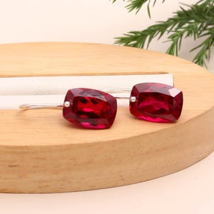 May include: A pair of silver earrings featuring large, faceted, rectangular, deep red gemstones. The earrings are displayed on a wooden surface with a neutral background and greenery.