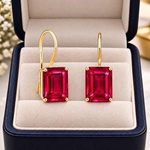 Può includere: Un paio di orecchini pendenti in oro con pietre preziose rosse dal taglio rettangolare a smeraldo. Ogni orecchino è sospeso a un gancio d'oro ed è presentato all'interno di un portagioie blu scuro con rivestimento beige.
