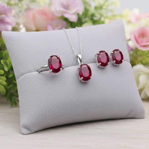 May include: A jewellery set comprising a ring, pendant necklace, and stud earrings. Each item features a silver setting and a large, oval-cut, deep red gemstone. The set is presented on a grey cushion with a floral backdrop.
